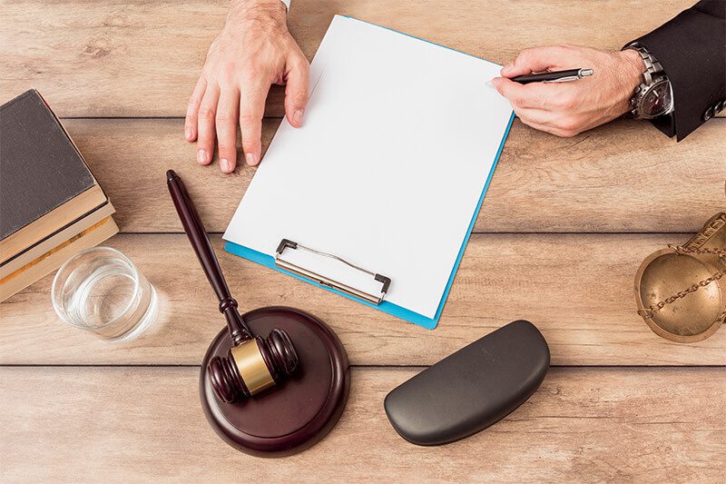A person begins to write on a blank piece of paper on a clipboard with a judge’s gavel, eyeglass case, and a glass of water within reach.